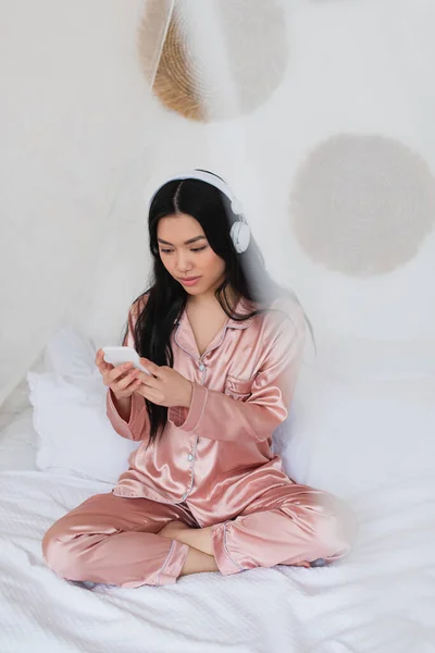 Mujer asiática joven en pijama de seda rosa sentada en la cama con auriculares y teléfono celular en el dormitorio - foto de stock