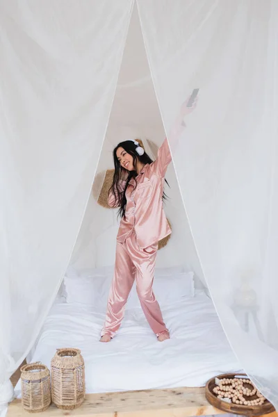 Positive young asian woman dancing on bed with headphones and holding cellphone with outstretched hand in bedroom — Stock Photo