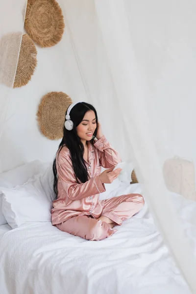 Positivo giovane donna asiatica seduta sul letto con le cuffie e guardando il cellulare in camera da letto — Foto stock