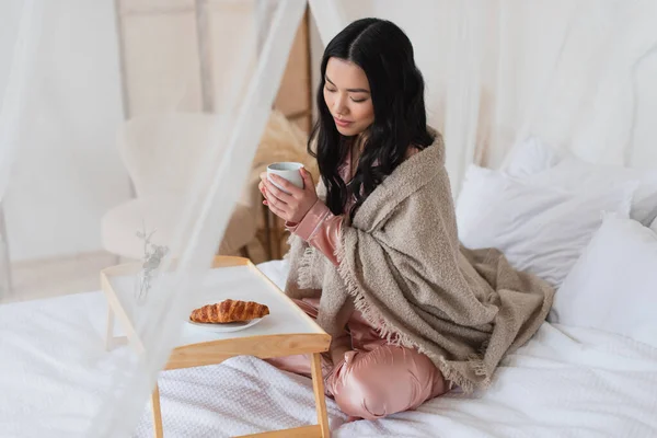 Young asian woman in silk pajamas and blanket sitting on bed with crossed legs and having breakfast in bedroom — Stock Photo
