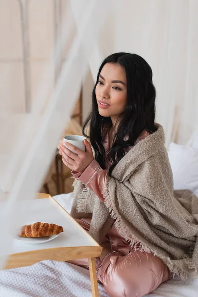 Молода азіатка в шовковій піжамі і ковдрі сидить на ліжку з чашкою кави біля столу з круасаном у спальні — стокове фото