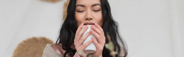 Porträt einer jungen Asiatin, die eine Tasse Kaffee in der Hand hält und Aroma im Schlafzimmer genießt, Banner — Stockfoto