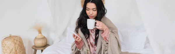 Young asian woman in silk pajamas and blanket sitting on bed, drinking coffee and holding cellphone in bedroom, banner — Stock Photo
