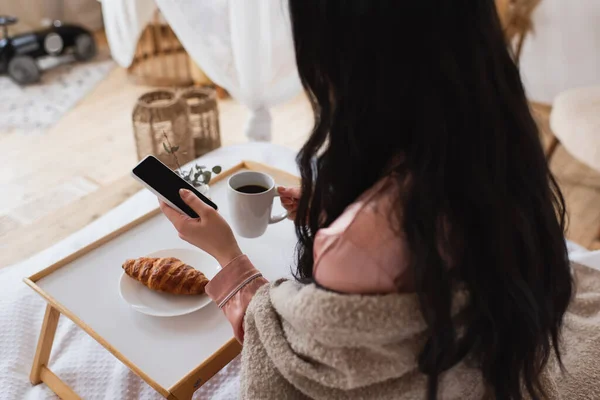 Teilansicht einer jungen brünetten Frau, die ihr Handy hält und auf dem Bett frühstückt — Stockfoto