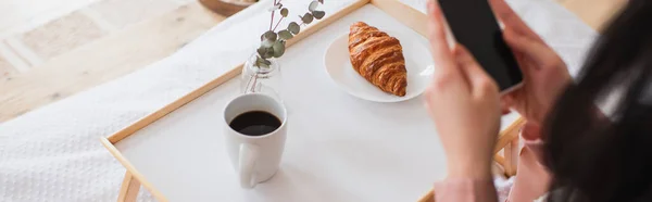 Cropped view of young brunette woman holding cellphone near table with coffee and croissant in bedroom, banner — Stock Photo