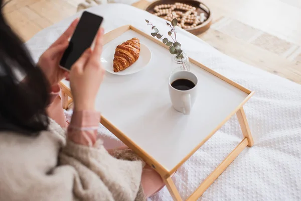 Частичный вид молодой брюнетки с телефоном рядом со столом с кофе и круассаном в спальне — стоковое фото