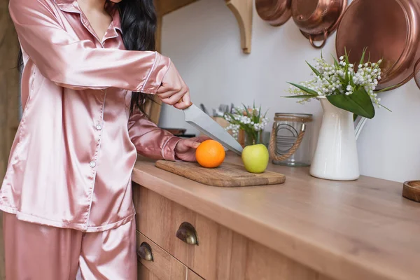 Partial view of young woman in pink silk pajamas cutting fruits in kitchen — Stock Photo