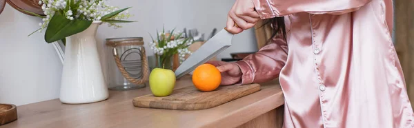 Vista recortada de mujer joven en pijama de seda rosa cortando frutas en la cocina, pancarta - foto de stock