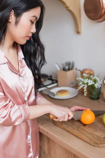 Junge Asiatin im rosa Seidenpyjama schneidet Orange in der Küche — Stockfoto