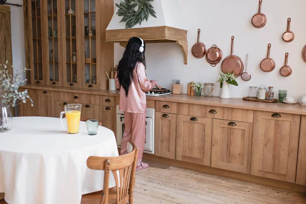Молодая азиатка в розовой шелковой пижаме и наушниках готовит завтрак на кухне — стоковое фото