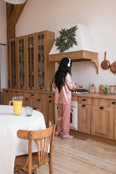 Jovem mulher asiática em pijama de seda rosa e fones de ouvido cozinhar café da manhã perto da mesa com suco de laranja na cozinha — Fotografia de Stock