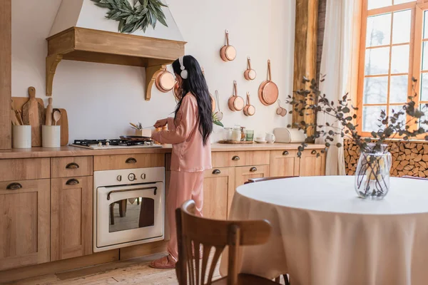 Young asian woman in pink silk pajamas and headphones cooking breakfast near table in kitchen — Stock Photo