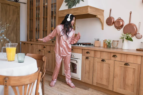 Joven asiático mujer en rosa seda pijama y auriculares cocinar desayuno y cantar con espátula en cocina - foto de stock