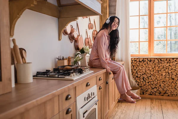 Sonriente joven asiática mujer en rosa pijama de seda y auriculares sentado cerca de la estufa y escuchar música en la cocina - foto de stock