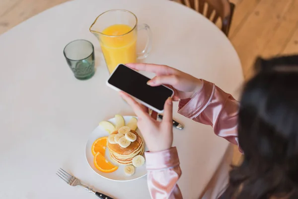 Partial view of young woman holding cellphone and taking picture of pancakes with fruits near orange juice in kitchen — Stock Photo