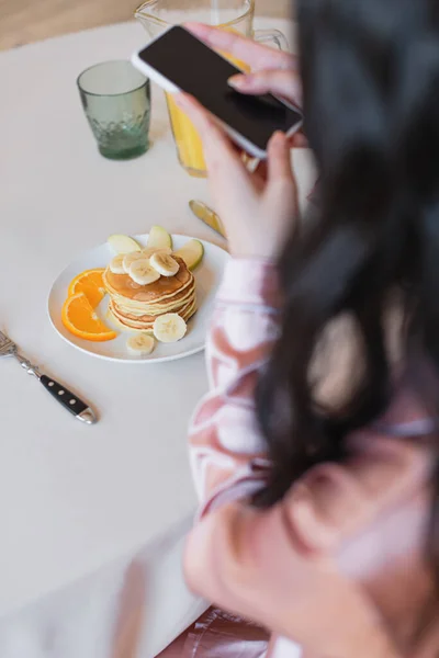 Vista cortada de jovem segurando celular e tirando foto de panquecas com frutas e suco de laranja na cozinha — Fotografia de Stock