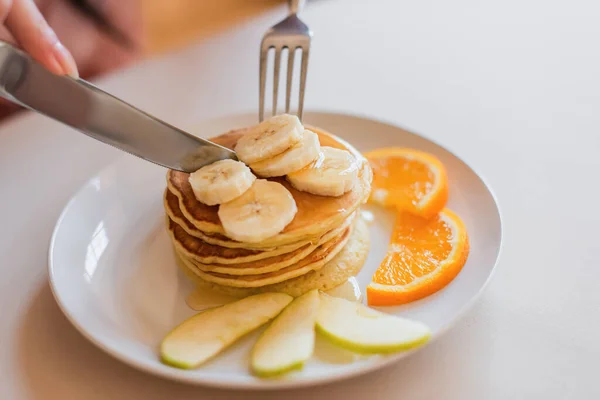 Vue rapprochée de fourchette en métal avec des crêpes coupantes au couteau avec des tranches d'orange et de banane sur plaque blanche dans la cuisine — Photo de stock