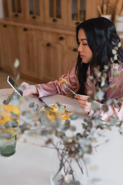 Позитивная молодая азиатка в шелковой пижаме ест блины с вилкой и смотрит на сотовый телефон с чистым экраном на кухне — стоковое фото