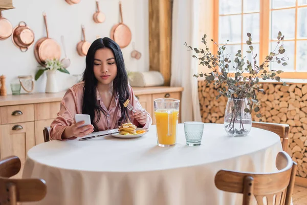 Lächelnde junge Asiatin im seidenen Pyjama, die auf dem Tisch sitzt und Pfannkuchen mit Gabel isst, während sie in der Küche auf das Handy schaut — Stockfoto