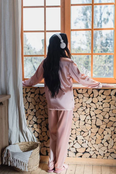 Backside view of young woman standing in headphones near window at home — Stock Photo