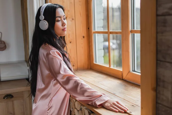 Soñadora joven asiática mujer en pijama de seda de pie cerca de la ventana con alféizar en los auriculares en casa - foto de stock
