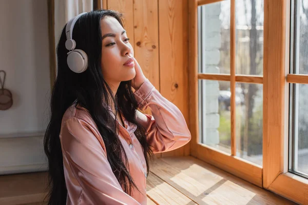 Mujer asiática joven soñadora en pijama de seda de pie cerca de la ventana con alféizar con auriculares y sosteniendo la mano cerca de la cabeza en casa - foto de stock