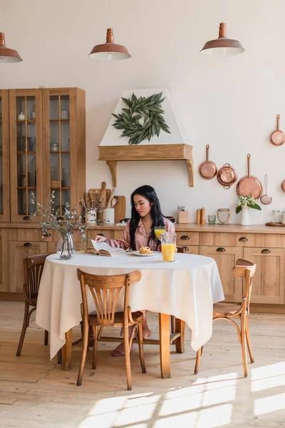 Jovem menina asiática em pijama de seda sentado na mesa e leitura livro enquanto tendo delicioso café da manhã na cozinha moderna — Fotografia de Stock