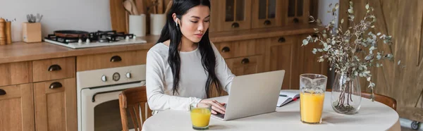 Serious young asian woman in earphones sitting on table and using laptop near notebook with pen in kitchen, banner — Stock Photo