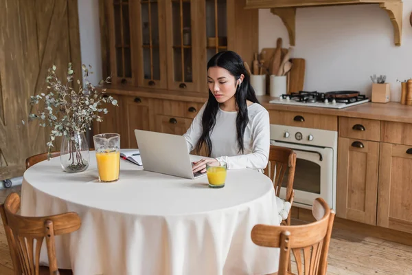 Serious young asian woman in earphones sitting on table and using laptop near notebook with pen in kitchen — Stock Photo