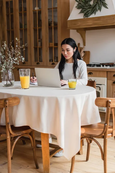 Seria joven asiática mujer en los auriculares sentado en la mesa, el uso de ordenador portátil y la escritura con pluma en el cuaderno en la cocina - foto de stock