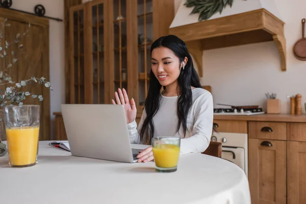 Jovem asiático mulher acenando mão e olhando para laptop enquanto tendo chamada de vídeo na cozinha — Fotografia de Stock