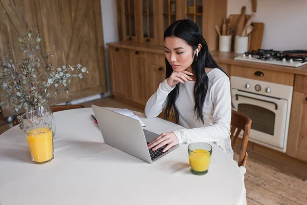 Jeune femme asiatique dans les écouteurs assis sur la table avec ordinateur portable et tenant la main près du visage dans la cuisine — Photo de stock