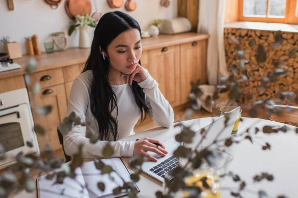 Young asian woman in earphones sitting on table and using laptop near notebook in kitchen — Stock Photo