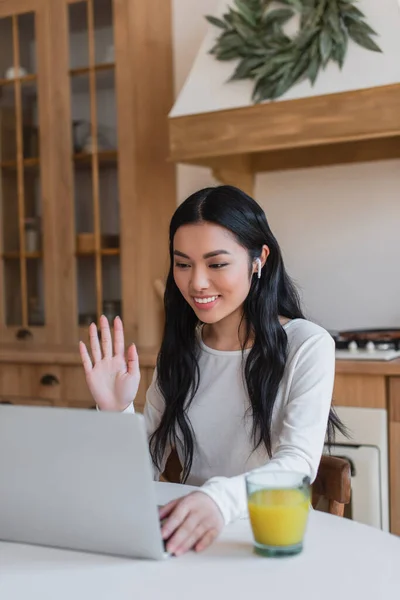 Sorridente giovane donna asiatica che agita la mano mentre fa videochiamata con laptop in cucina — Foto stock