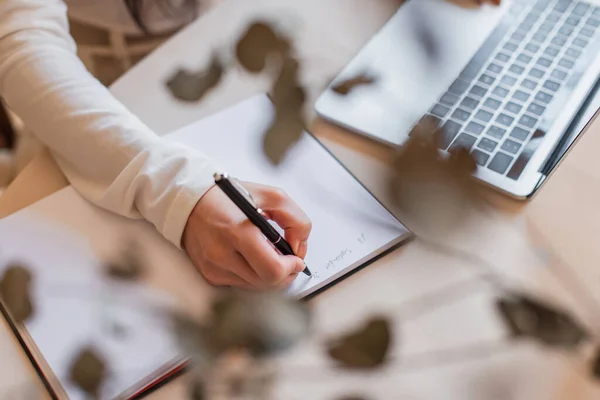 Vista parcial de la escritura femenina joven en el cuaderno cerca de la computadora portátil en la mesa - foto de stock