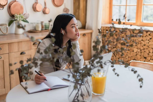 Dreamy young asian woman in earphones sitting on table with pen, notebook and laptop in kitchen — Stock Photo
