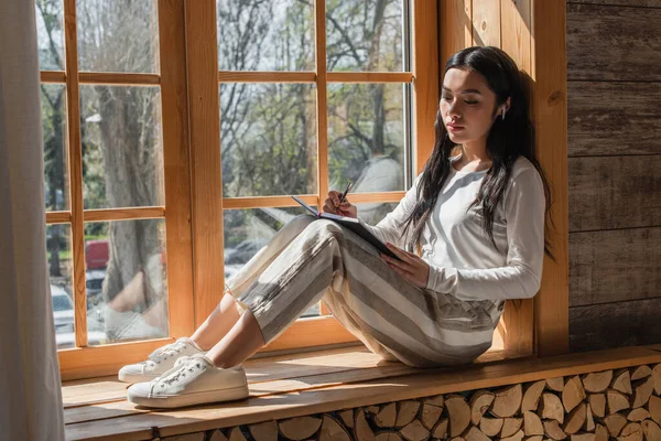 Мечтательная молодая азиатка в повседневной одежде и наушниках сидит у окна с ручкой и ноутбуком дома — стоковое фото
