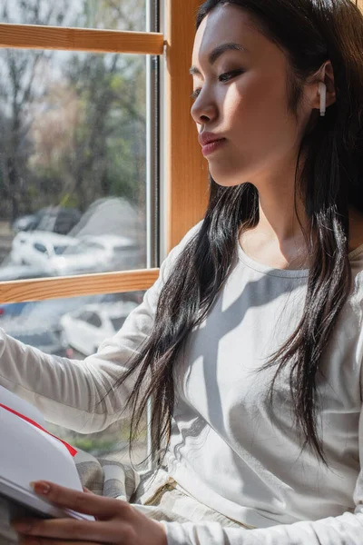 Sognante giovane donna asiatica in auricolare seduta vicino alla finestra con notebook a casa — Foto stock