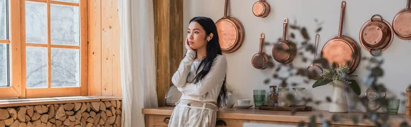 Nachdenkliche junge asiatische Frau mit Kopfhörern, die mit der Hand vor dem Gesicht in der Küche steht, Banner — Stockfoto
