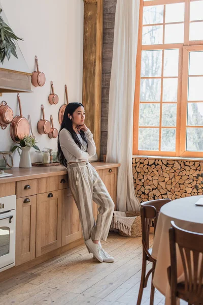 Thoughtful young asian woman in earphones standing with hand near face in kitchen — Stock Photo