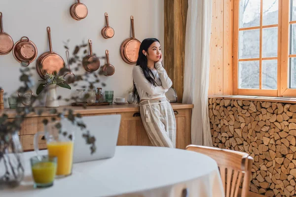 Pensativo jovem asiático mulher em blusa e calças de pé com fones de ouvido e segurando a mão perto do rosto na cozinha — Fotografia de Stock