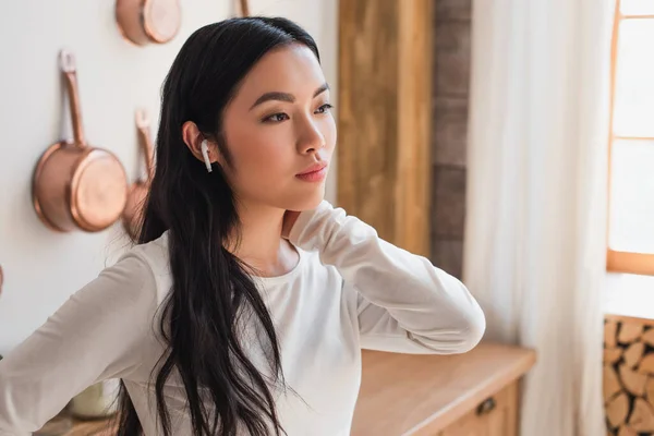 Cansado joven asiático mujer en blanco blusa de pie con auriculares y celebración de la mano cerca del cuello en la cocina - foto de stock