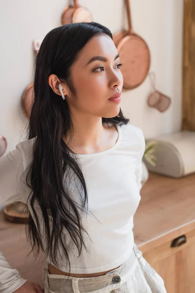 Portrait of serious young asian woman in white blouse standing with earphones in kitchen — Stock Photo