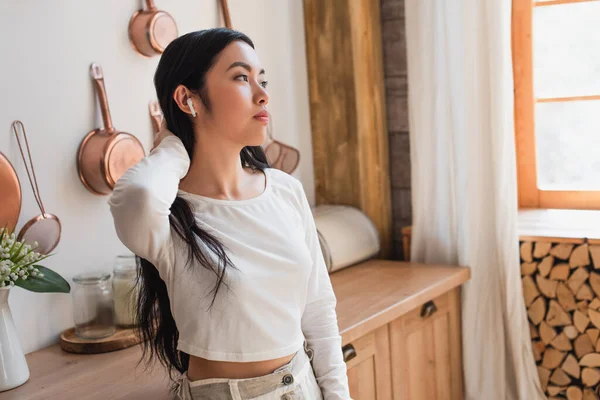 Tired young asian woman in white blouse standing with earphones and holding hand near neck in kitchen — Stock Photo