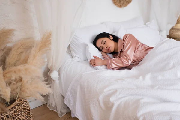 Pretty young asian woman in silk pajamas sleeping in bed with white linen in modern bedroom — Stock Photo