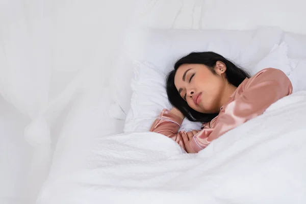 Jeune femme asiatique en pyjama de soie dormant dans le lit avec du linge blanc dans la chambre — Photo de stock