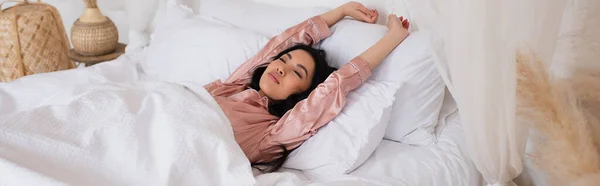 Joven mujer asiática en pijama de seda despertando con las manos extendidas y los ojos cerrados en el dormitorio, pancarta - foto de stock