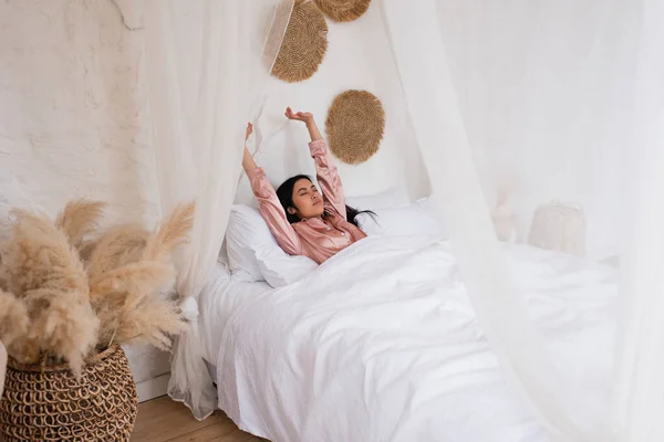 Somnoliento joven asiático mujer en pijama de seda despertando con las manos extendidas en el dormitorio - foto de stock