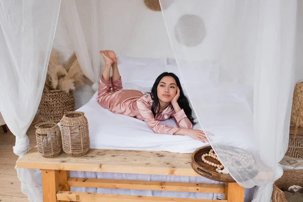 Jeune femme asiatique en pyjama de soie couché avec la main près du visage sur du linge blanc dans la chambre — Photo de stock