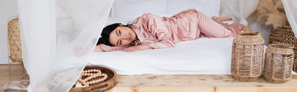 Young asian woman in silk pajamas lying with outstretched hand on white linen in bedroom, banner — Stock Photo